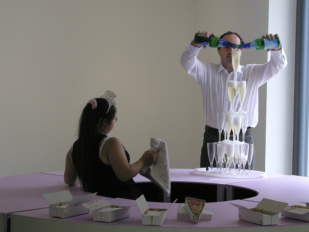 A photograph of dancer Aisha Booth and artist Charlie Fox. Charlie is pouring champagne into a fountain of glasses and Aisha leans back to watch. They are standing around a large round wooden table that has been created to look like a cake. They are at Café Gallery Projects gallery, now called Southwark Park Galleries.